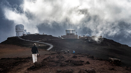 Observatory buildings on a rocky hill under dramatic clouds with people walking in the scene. Kula, Hawaii, USA