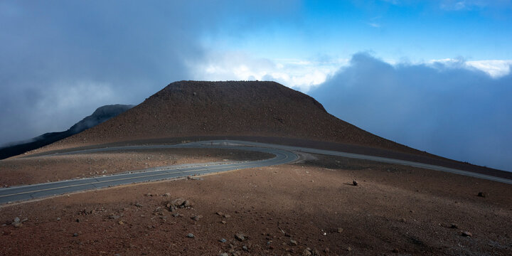 A winding road curves around a barren, reddish-brown mountain under a bright, cloudy sky. Kula, Hawaii, USA