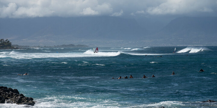 Surfers and windsurfers navigate waves against a backdrop of mountains under a cloudy sky. Haiku, Hawaii, USA