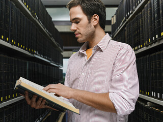 A young adult reading a book in a library aisle surrounded by shelves of dark books. Germany