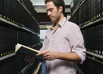 Young man reading a book in a library aisle surrounded by shelves of black hardcover books. Germany