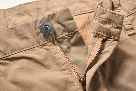 Close-up of beige pants, metal button and zipper partially unzipped, showing stitching details.
