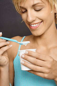 Smiling woman in a blue top enjoying yogurt with a spoon from a white container. Germany