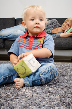 Toddler playing with a soft block on a fuzzy rug while a woman relaxes on a couch in the background. Germany