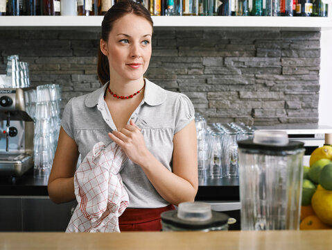 Woman in a caf&Egrave; drying a glass behind the counter with a neutral expression. Germany