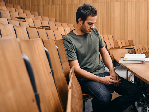 Young man seated in an empty lecture hall, focused on his phone with books beside him. Germany