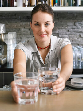 Woman in a polo shirt offering glasses of water with a smile in a modern kitchen. Germany