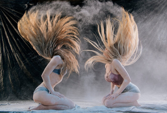 Two women kneel with hair flipping amid dramatic powder clouds against a dark backdrop. WA, USA