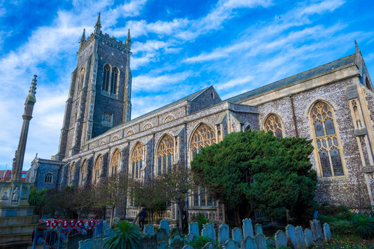 Historic stone cathedral under a vibrant blue sky with surrounding trees and gravestones. Norfolk,Cromer, England