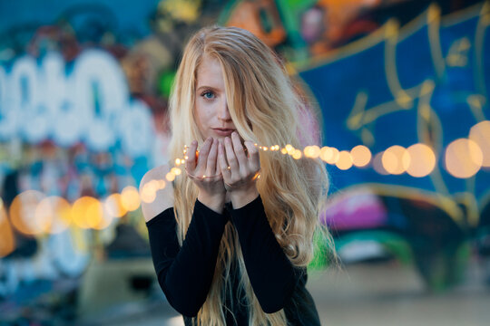 Woman with long blonde hair holds string lights in front of colorful graffiti backdrop WA, USA