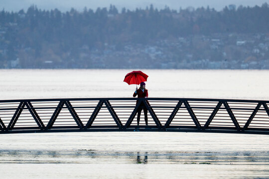 Person with red umbrella standing on a bridge over calm water with forested background. Bainbridge Island, WA, USA