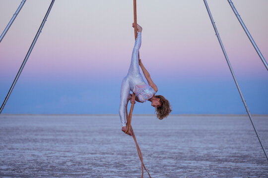 Aerial performer in white costume gracefully hangs upside down on outdoor rig at dusk. WA, USA