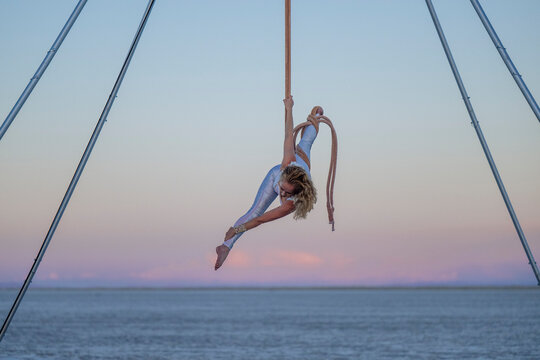 Aerial performer in silver outfit skillfully hangs on ropes over the ocean at sunset. WA, USA