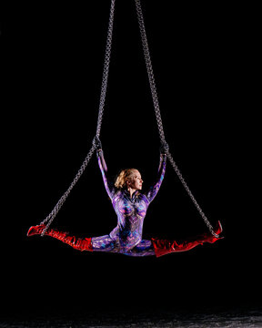 Female aerialist in splits on chains in colorful bodysuit and red boots against black background. WA, USA