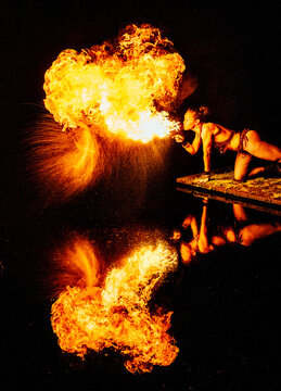A performer breathes fire, creating a dramatic reflection on the dark water surface. WA, USA