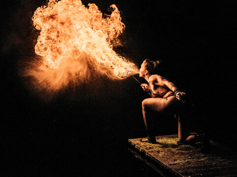 Fire performer kneeling on wooden platform, breathing a dramatic burst of flames into the night. WA, USA