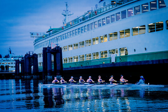 Rowing team practices on calm water near a large ferry at dusk. WA, USA