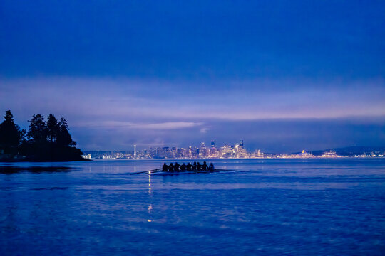 Rowers glide across a tranquil lake with a city skyline glowing under a deep blue evening sky. WA, USA