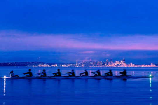 Eight rowers in a boat with a blurred city skyline under a glowing twilight sky. WA, USA