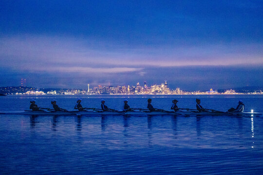 Eight rowers scull across calm waters at dusk with a city skyline in the background. WA, USA