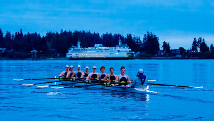 Rowing team practices on a calm lake with a ferry and forested background under a blue sky. WA, USA