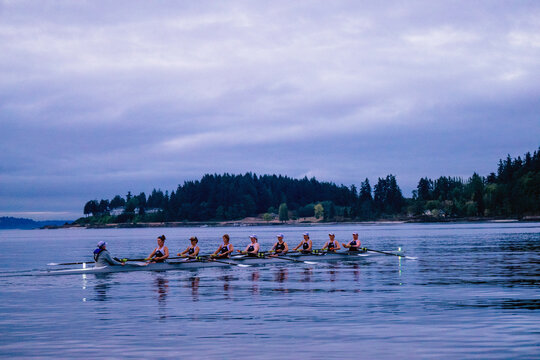 Rowing team glides through calm waters under cloudy skies with a forested shoreline backdrop. WA, USA