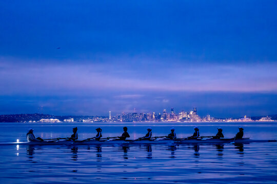 Rowing team practices at dawn with city skyline in the background over calm water. WA, USA