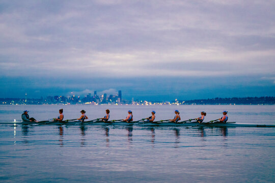 Rowers glide on calm water with a city skyline and cloudy sky in the background. WA, USA