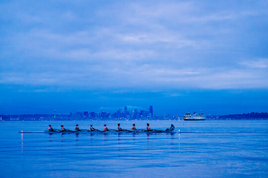A rowing team glides across calm water with a cityscape and ferry in the distance at dusk. WA, USA