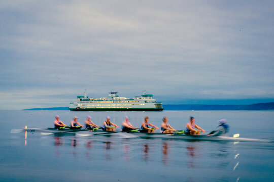 A rowing team glides on water with a ferry in the background under a cloudy sky. WA, USA