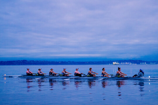 Rowing team practicing on calm water at dusk under a cloudy sky with distant city lights. WA, USA