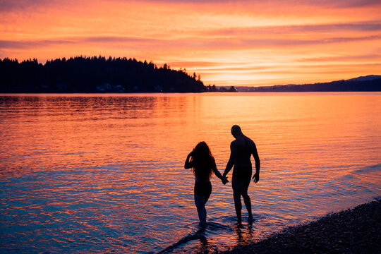 A couple walks hand in hand along the shore during a vibrant sunset over calm waters. WA, USA
