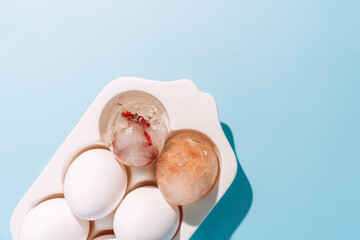Egg carton with white eggs and decorative ice egg on blue background, minimal still life. High quality photo