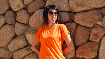 woman in orange dress standing against rustic stone wall, wearing sunglasses outdoors.