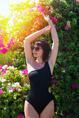 Confident Woman in Black Swimsuit Posing Gracefully Against a Backdrop of Vibrant Flowering Bushes in Lush Garden Setting.