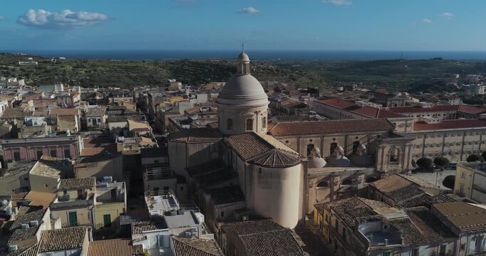 Aerial view of the majestic Noto Cathedral, its dome piercing the skyline amidst a sea of terracotta rooftops, bathed in the warm glow, Noto, Sicily, Italy.