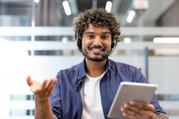 Portrait of a young smiling Indian man in a headset sitting at a desk in the office in front of the camera and talking on a video call