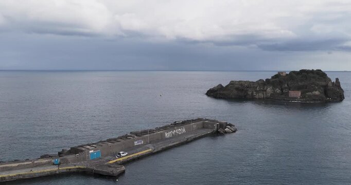 Aerial view of the dark blue waters surrounding the rocky Lachea Island near a concrete pier under a cloudy sky, Aci Trezza, Sicily, Italy.