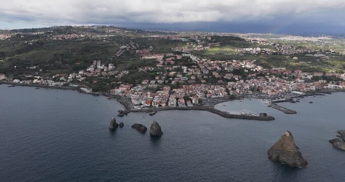 Aerial view of Aci Trezza and the imposing Faraglioni rocks emerging from the deep blue sea, a picturesque contrast against the coastal town, Aci Trezza, Sicily, Italy.