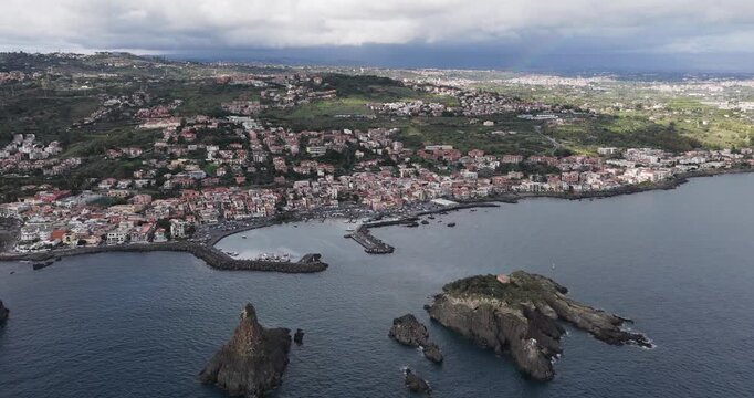 Aerial view of the coast with the distinctive Faraglioni rocks contrasting against the dark sea and the town's buildings, Aci Trezza, Sicily, Italy.