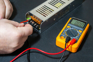 hands using a yellow digital multimeter with red and black probes to test the terminals of a metal power supply unit featuring a lit green LED indicator.
