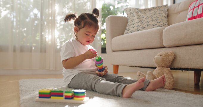 A two year old Asian girl sits barefoot on a soft gray rug, stacking ring toys near sheer curtains, a beige sofa, patterned pillows, and a plush teddy bear.
