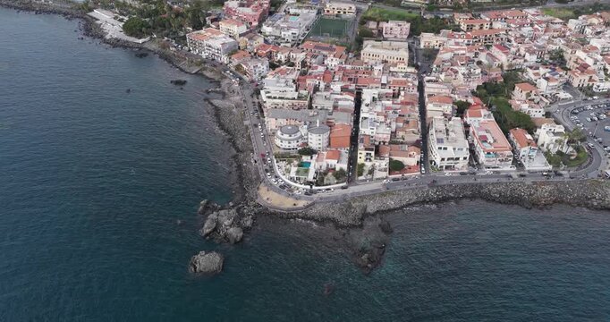 Aerial view of the dark blue sea meeting the rocky shore and vibrant buildings of Aci Trezza, a coastal town with a bustling promenade, Aci Trezza, Sicily, Italy.