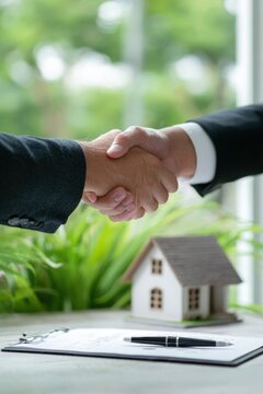 Real estate business market, house home building purchase sale. Two men shaking hands over a clipboard with a house model on it, surrounded by lush green plants and a blurred background.
