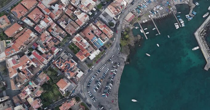 Aerial view of vibrant red-roofed buildings contrast against the turquoise sea and bustling port, showcasing Aci Trezza's coastal charm, Aci Trezza, Sicily, Italy.