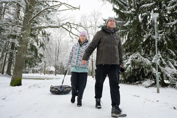 Man and a woman walk through a snowy winter park holding hands and pulling a inflatable donut sled