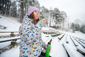 Girl drinks a hot drink from a thermos mug in a snowy winter park.