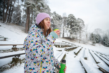 Girl drinks a hot drink from a thermos mug in a snowy winter park