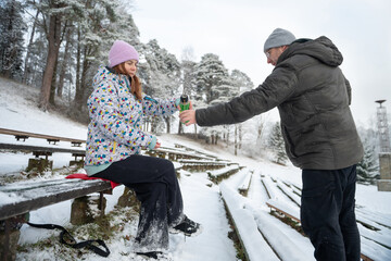 Woman pours a hot drink from a thermos into a man's mug in a snowy winter park