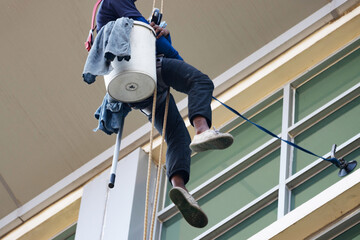 Professional rope access window cleaner working at height on building exterior using safety harness and bucket, high-rise maintenance service concept
